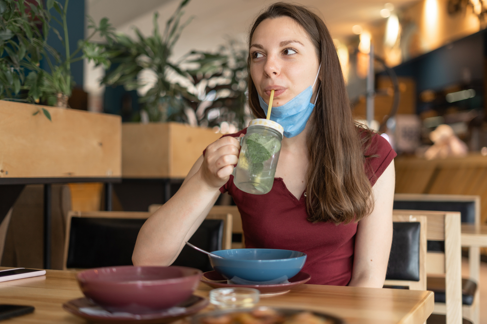 A woman is drinking through a straw in a restaurant with her mask pulled under her chin.